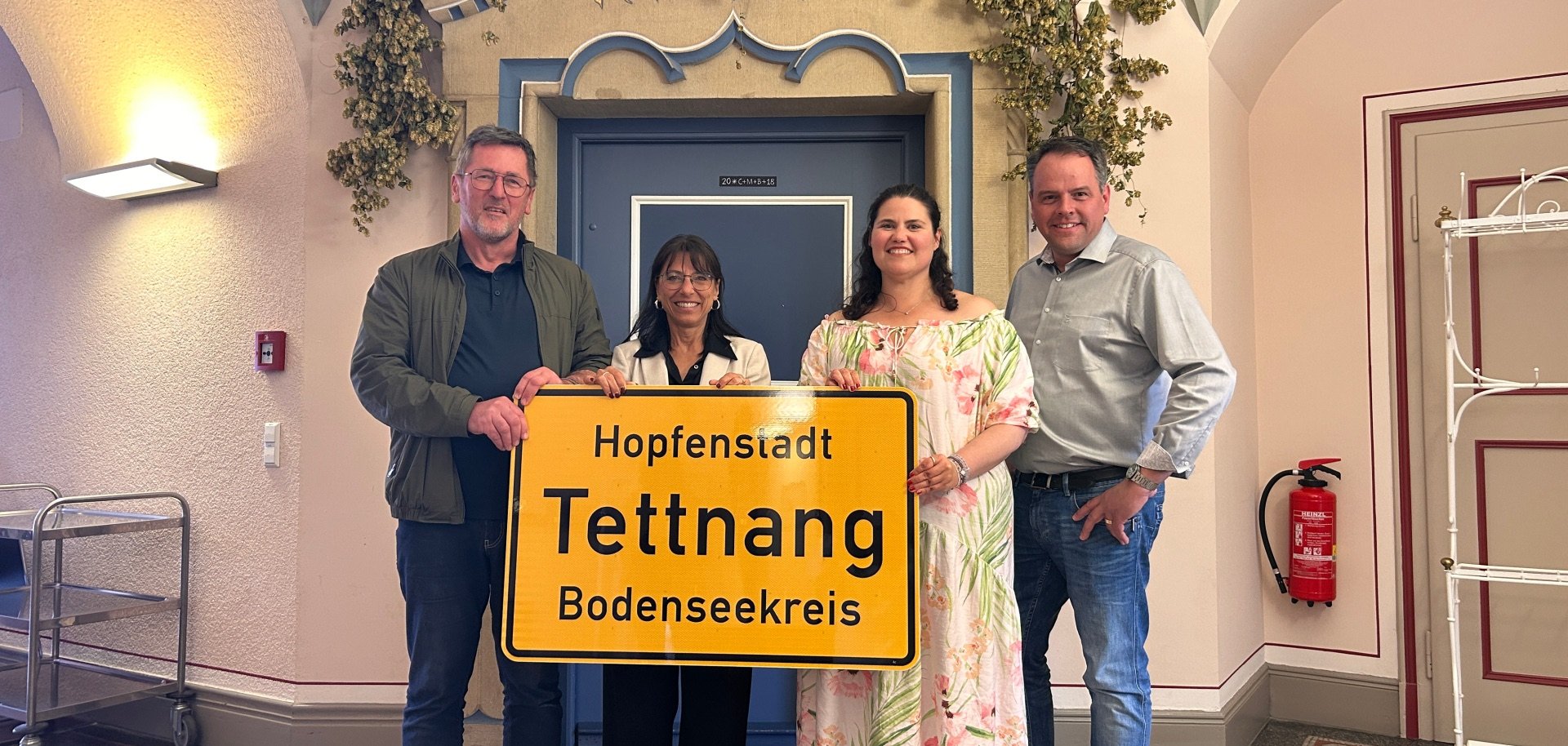 Vier Personen vor dem Sitzungssaal des Rathauses tragen das neue Ortsschild mit dem Namenszusatz Hopfenstadt 