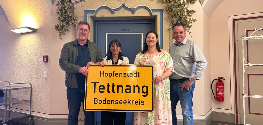 Vier Personen vor dem Sitzungssaal des Rathauses tragen das neue Ortsschild mit dem Namenszusatz Hopfenstadt 