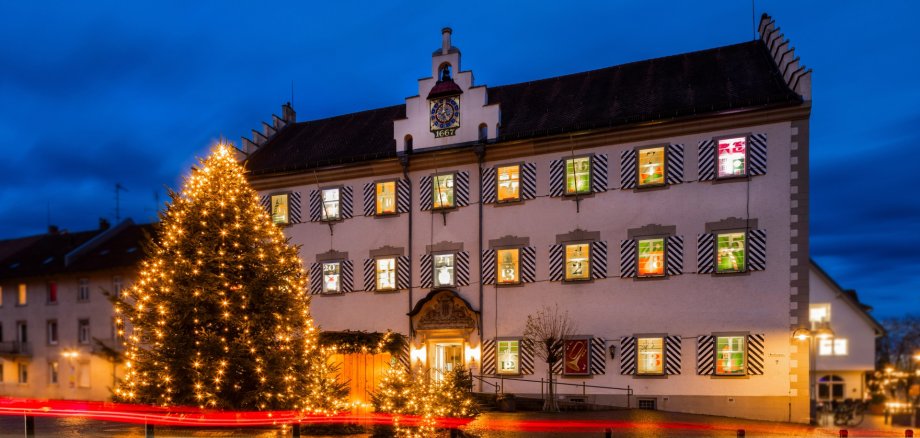 Vorderansicht des Tettnanger Rathauses mit den beleuchteten Fenstern des Adventskalenders. Im Vordergrund der beleuchtete Weihnachtsbaum und das Himmlische Postamt.